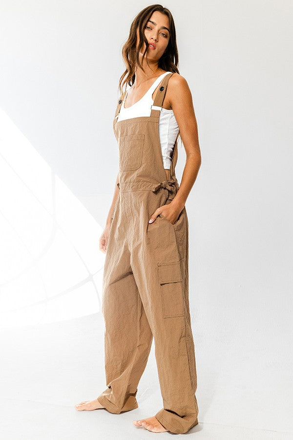 woman wearing a tan linen overall with adjustable straps and side pockets, styled with a white tank top underneath