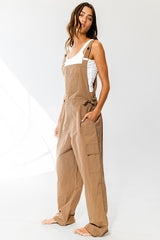woman wearing a tan linen overall with adjustable straps and side pockets, styled with a white tank top underneath