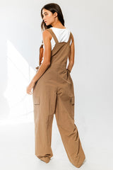 woman wearing a back view of a brown utility jumpsuit with wide legs and adjustable straps over a white tank top