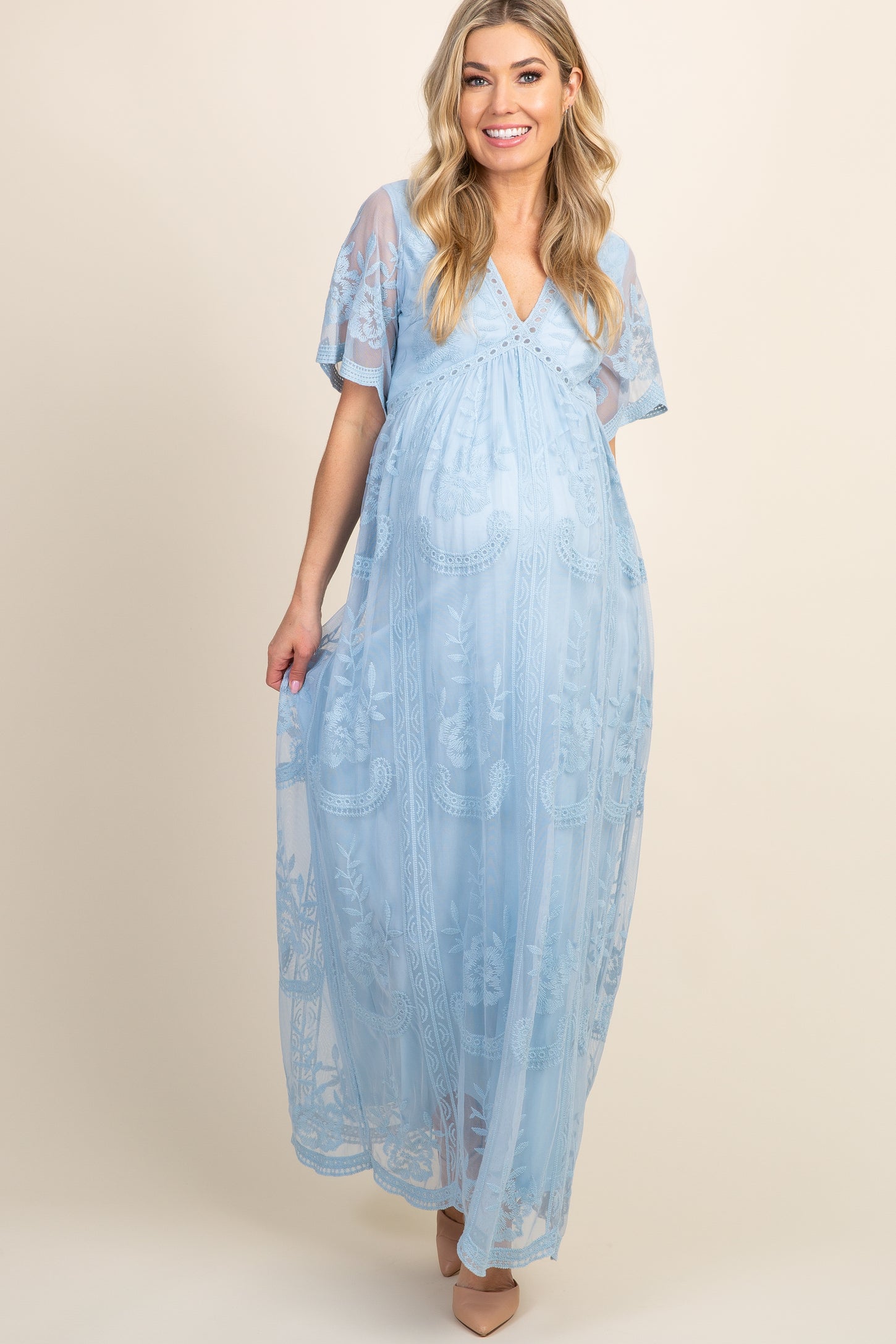 woman wearing a light blue maternity maxi dress with floral embroidery and a visible baby bump, standing in front of colorful balloons