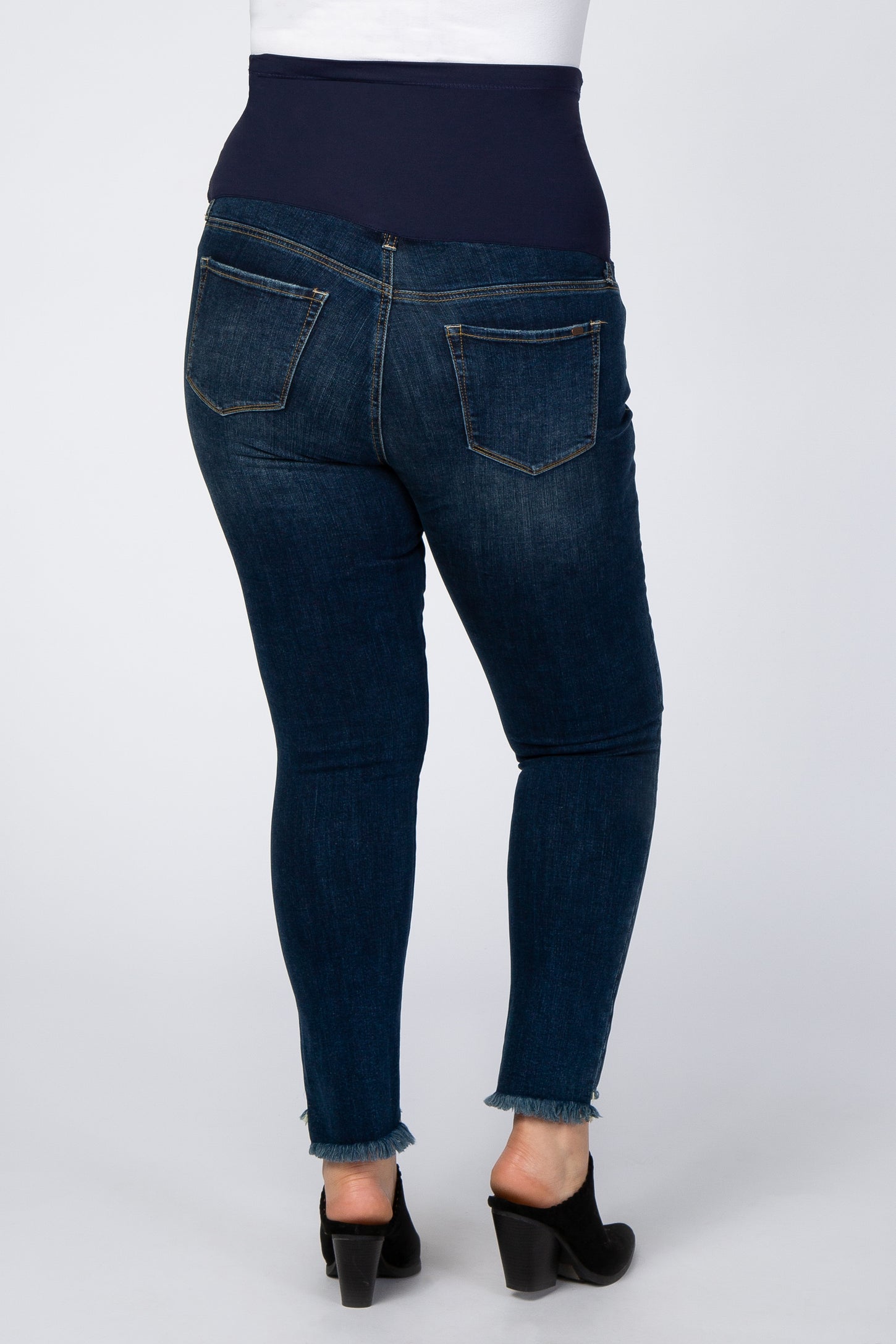 Back view of dark wash maternity skinny jeans with a belly band and frayed hems on a white background