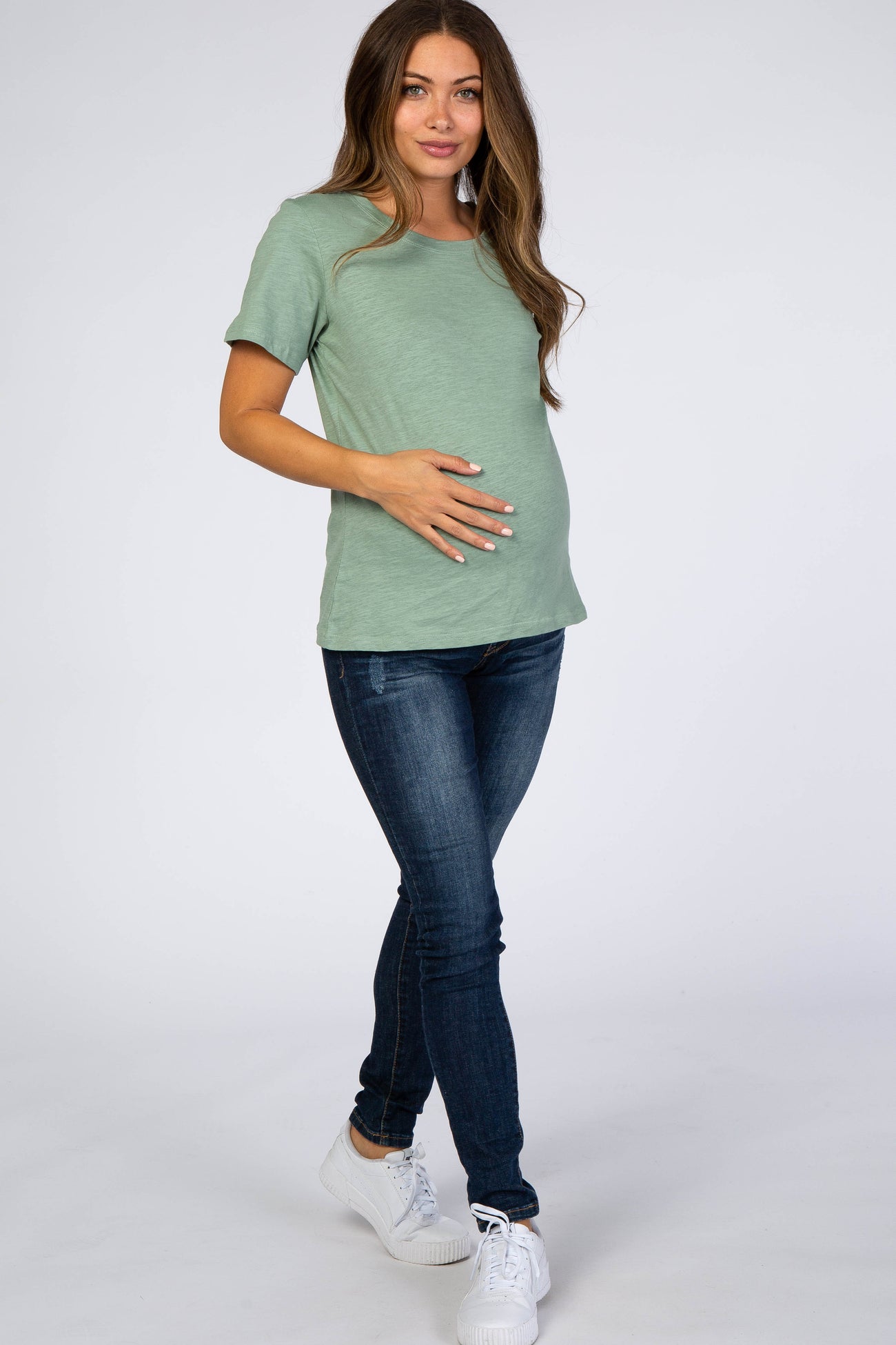 pregnant woman in sage green maternity t-shirt and dark wash jeans posing against a white background