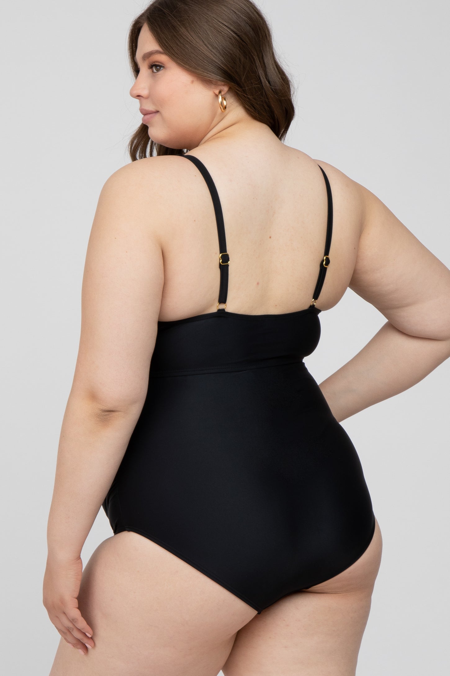 Back view of a woman wearing a plus size black one-piece swimsuit with adjustable gold hardware straps