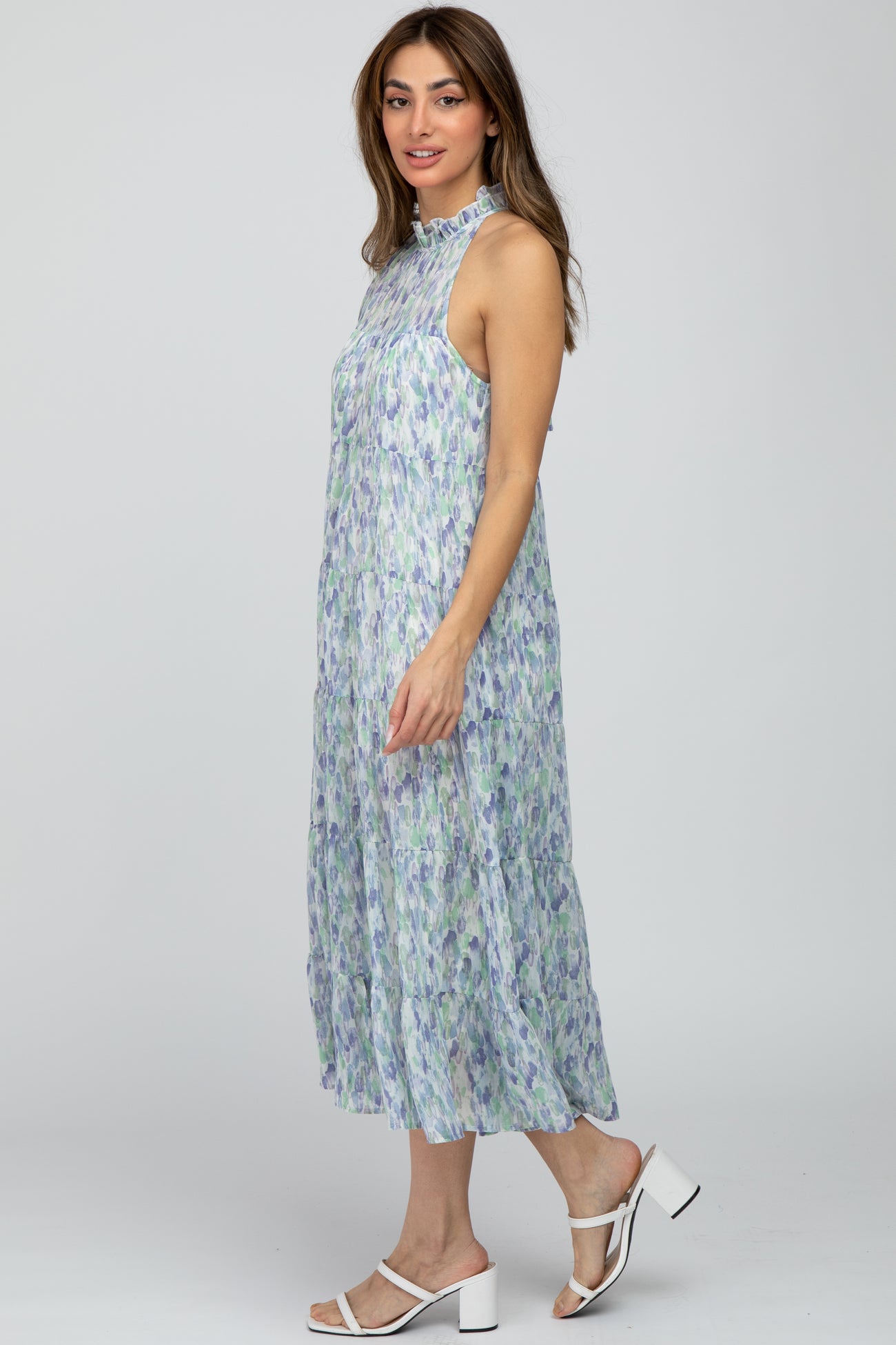 woman wearing a flowy sleeveless midi dress with a floral pattern in green and purple, paired with white block heel sandals
