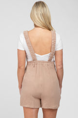 Back view of a woman wearing tan gauze overalls over a white t-shirt against a grey background