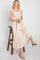 woman in cream smocked midi dress with ruffle straps sitting on a bamboo stool holding a straw handbag