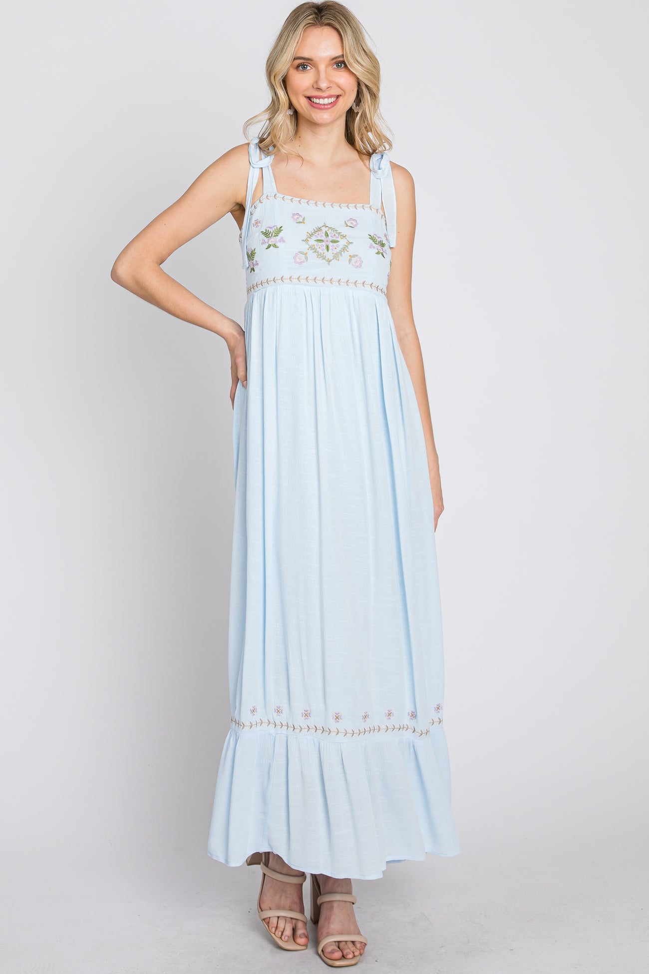 Woman wearing a light blue maxi dress with floral embroidery and tie straps against a white background