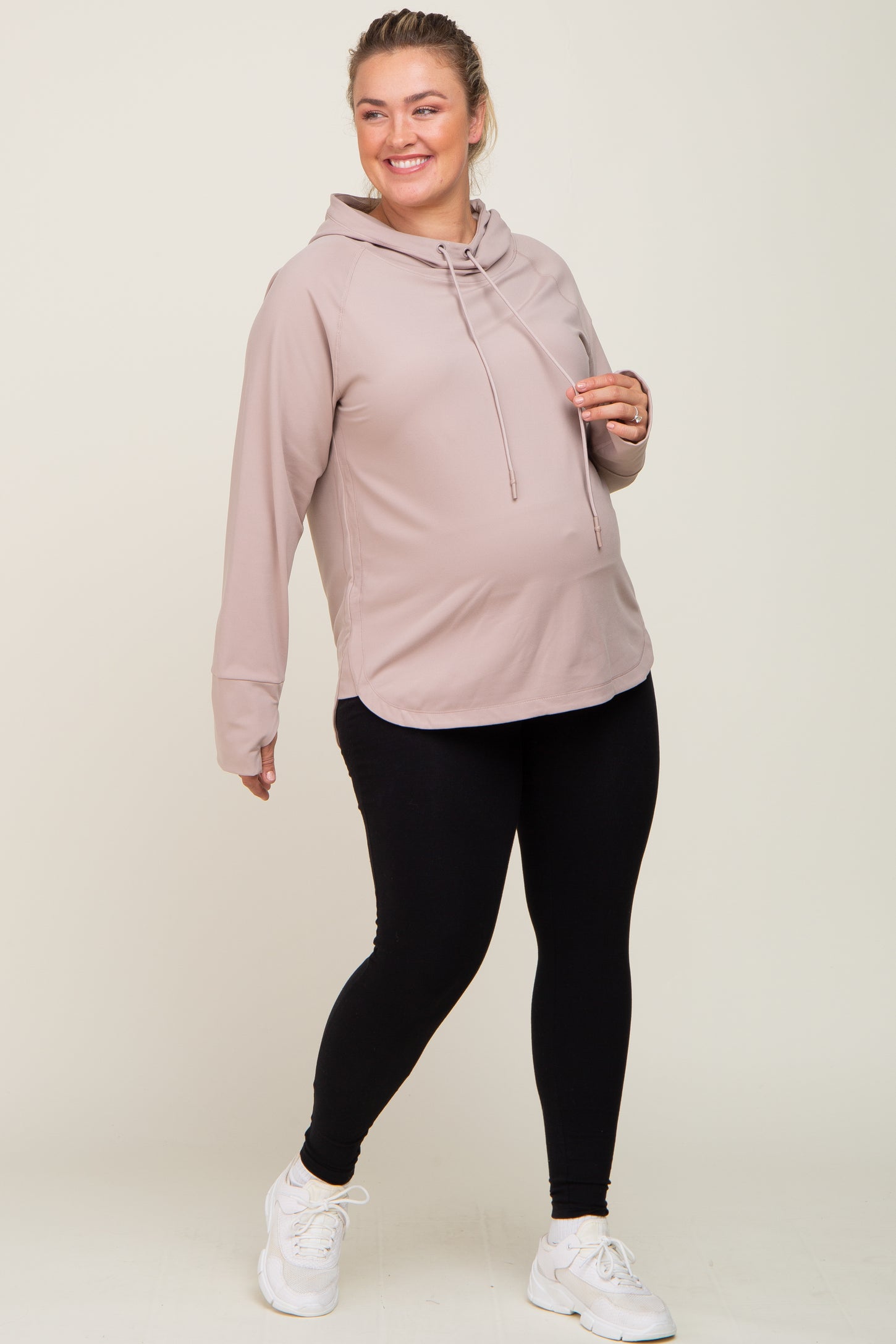 Smiling pregnant woman wearing a mauve maternity hoodie and black leggings with white sneakers