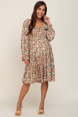 Smiling woman wearing a long sleeve floral smocked maternity dress with tiered skirt and tan heels