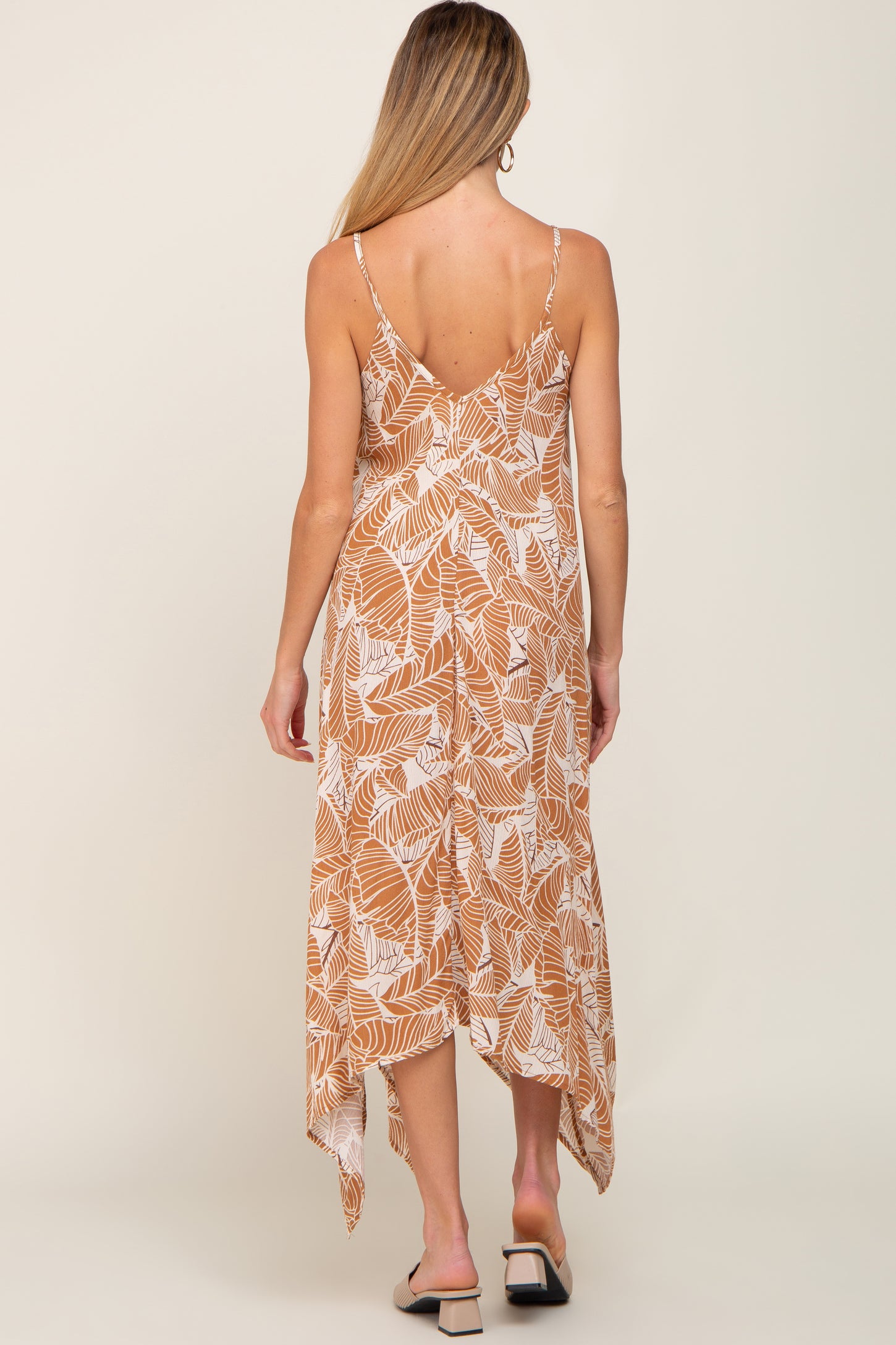 Back view of a woman wearing a tan and white leaf print midi dress with spaghetti straps and handkerchief hem