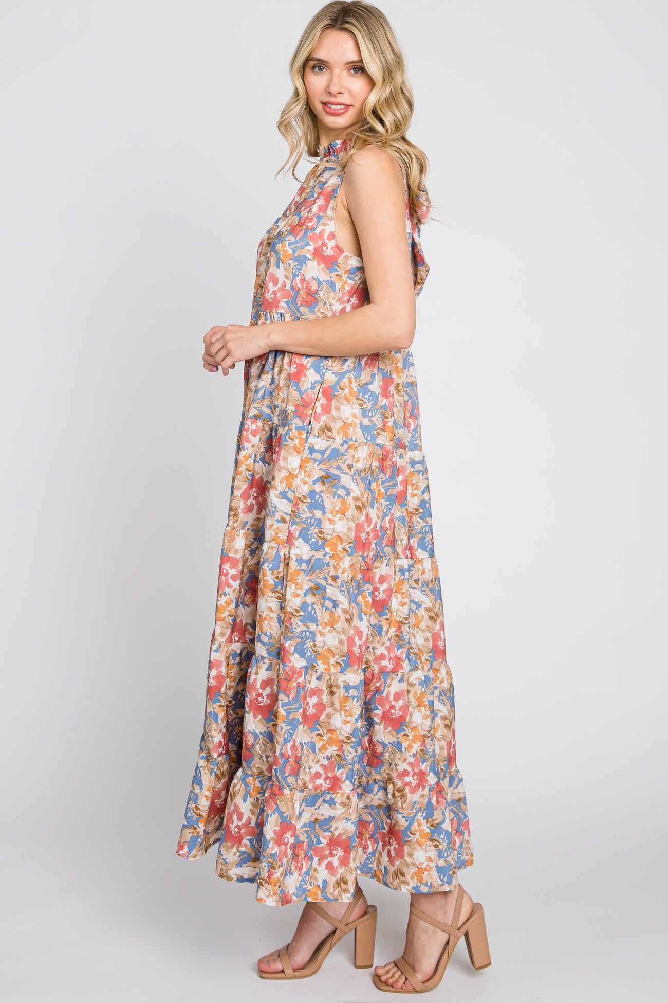 Woman wearing a sleeveless floral tiered maxi dress and tan block heel sandals against a white background