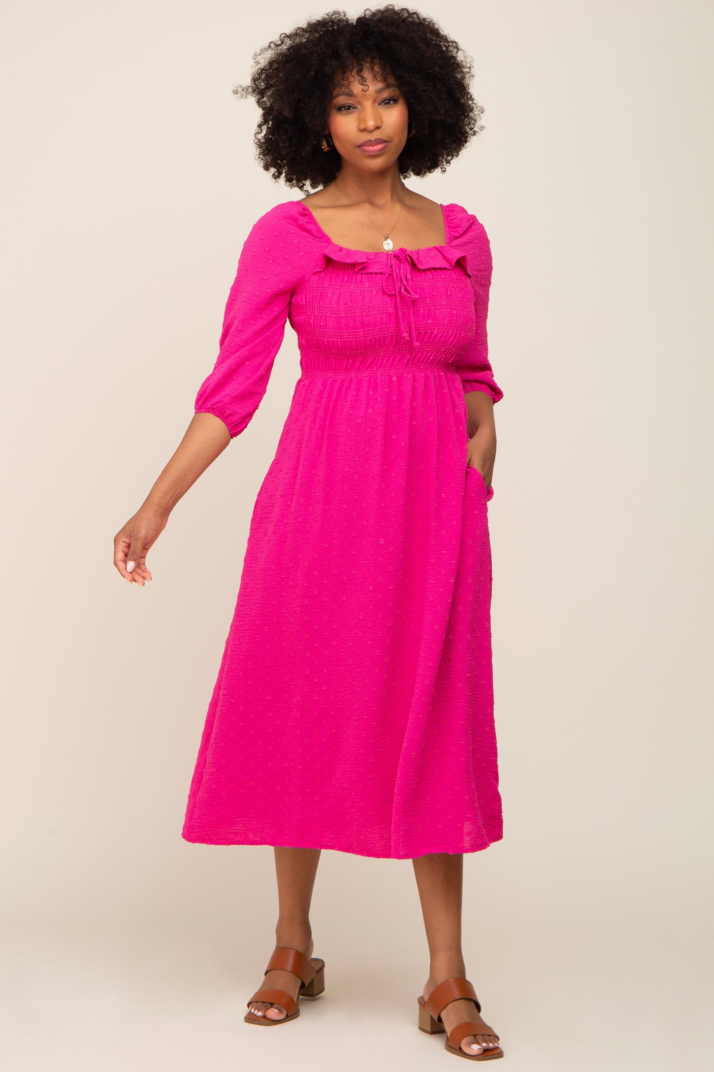 woman wearing a bright pink midi dress with puff sleeves and a tie neckline, posing with one hand in pocket