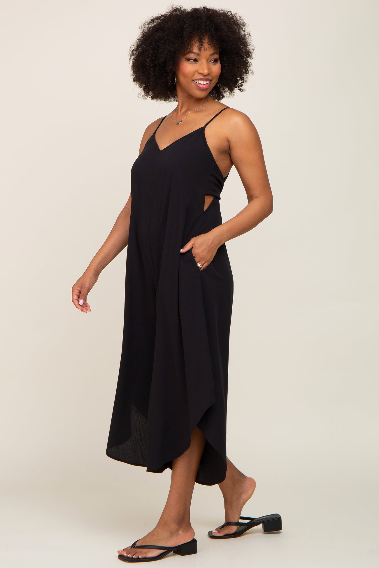 Woman with afro hair wearing a black sleeveless maternity jumpsuit with side cutouts and pockets