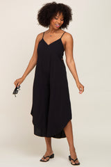 Woman with afro hair wearing a black sleeveless wide-leg jumpsuit and holding sunglasses against a plain background