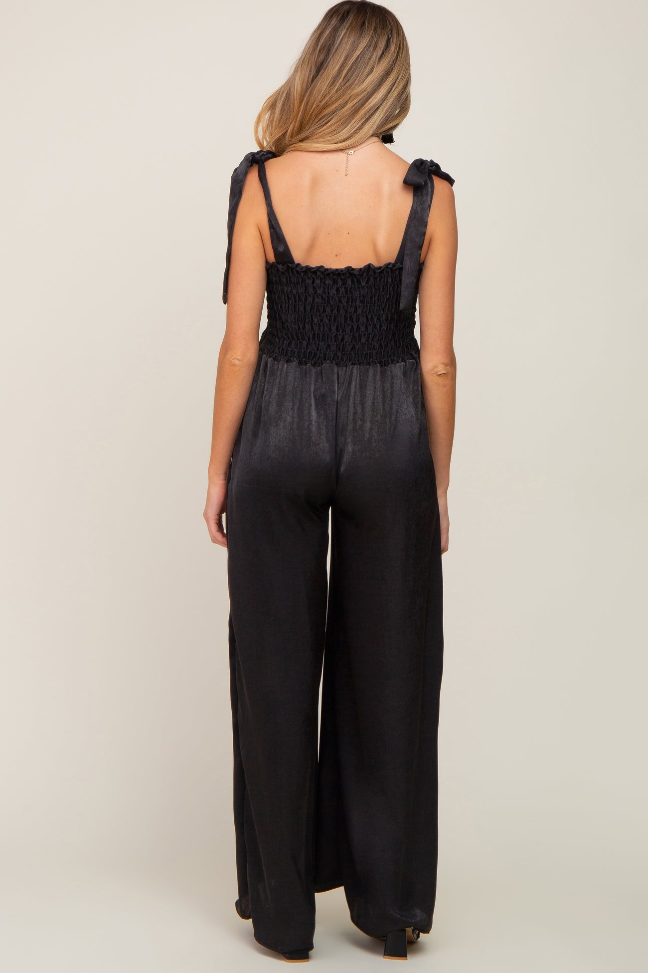 Back view of a woman wearing a black satin jumpsuit with smocked bodice and tie straps