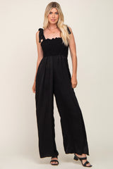 Woman wearing a black smocked wide leg jumpsuit with tie straps and block heel sandals