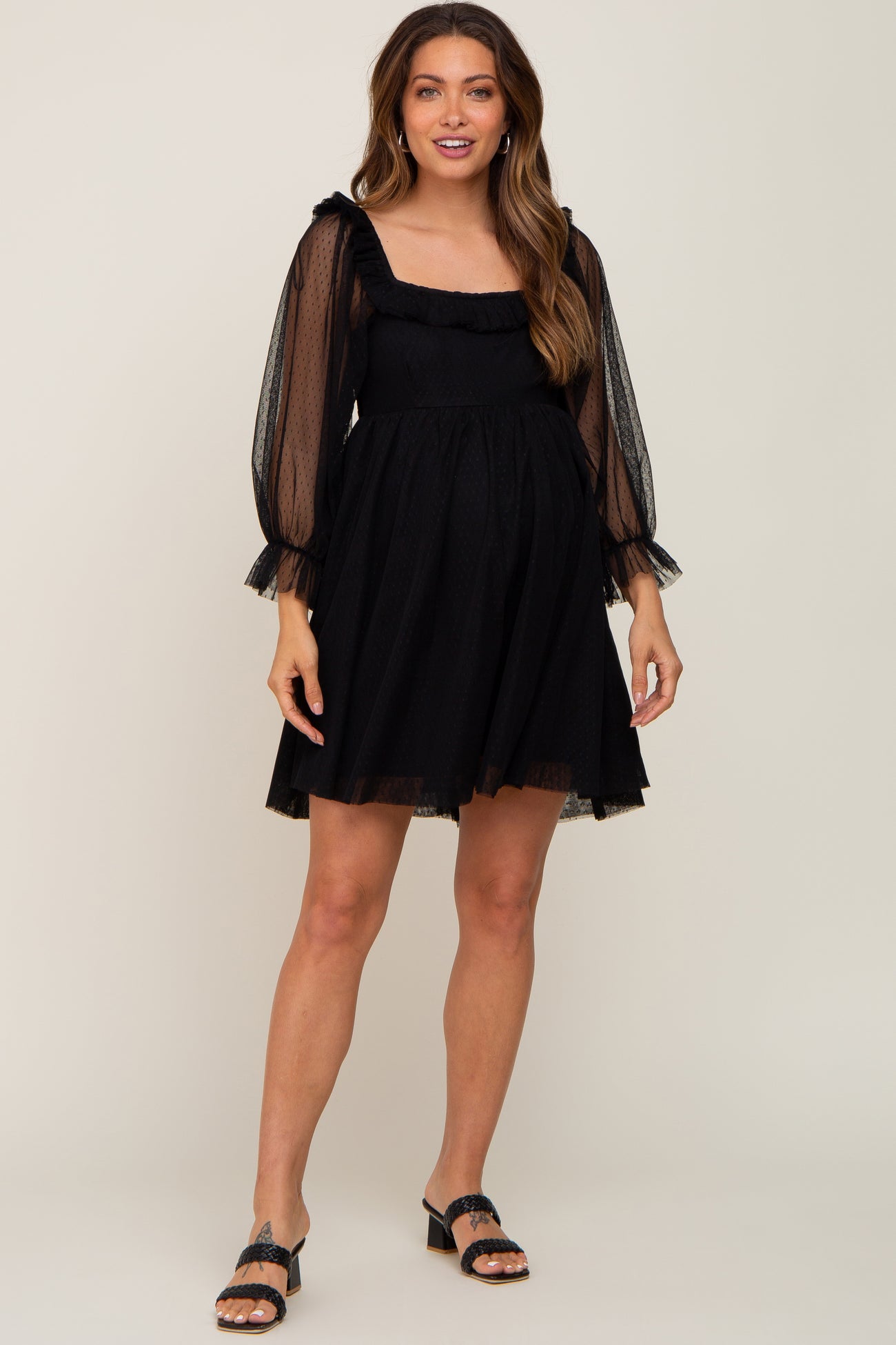 Pregnant woman wearing a black mesh long sleeve maternity mini dress with ruffle details and black heels