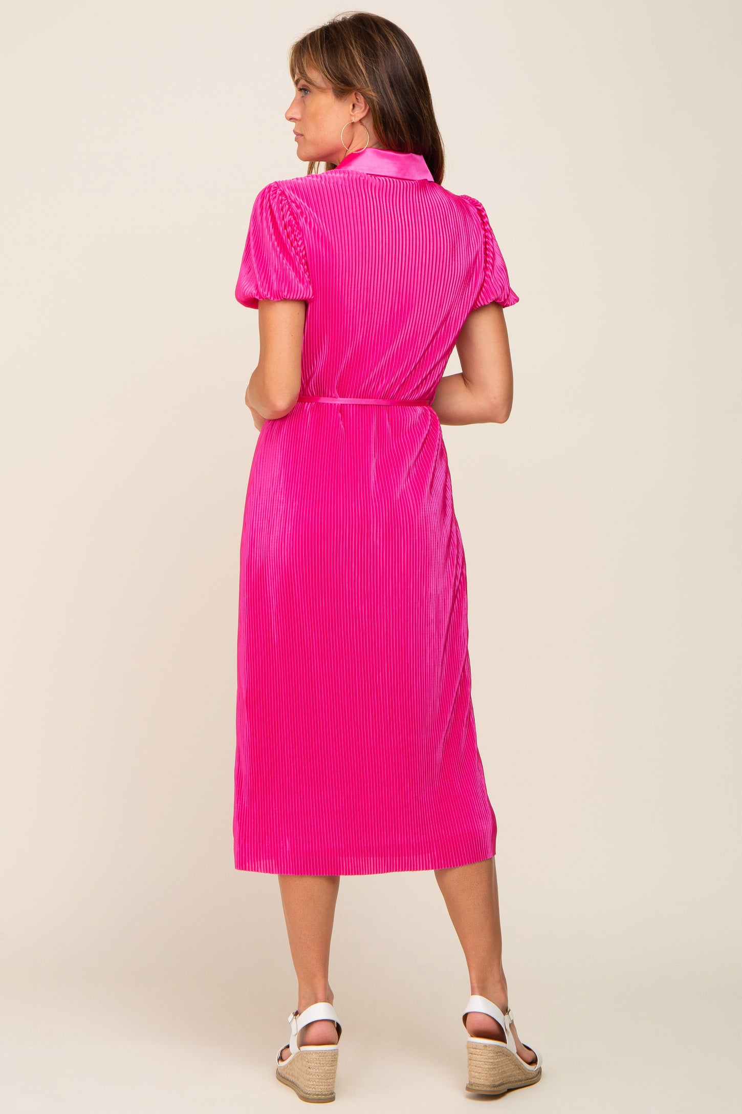 Back view of a woman wearing a hot pink pleated midi dress with short sleeves and white wedge sandals