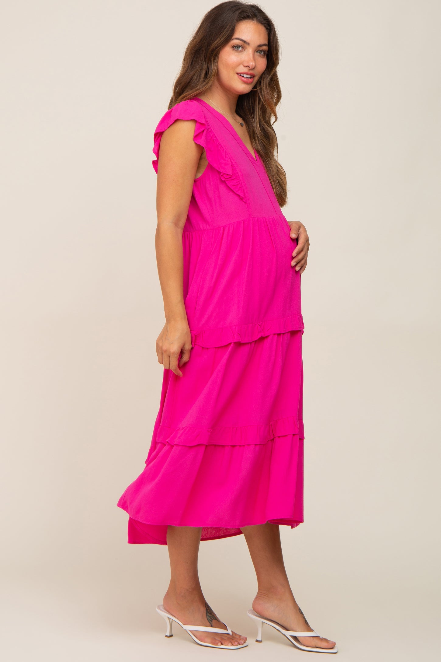 Pregnant woman wearing a hot pink ruffled tiered maternity midi dress and white kitten heel sandals