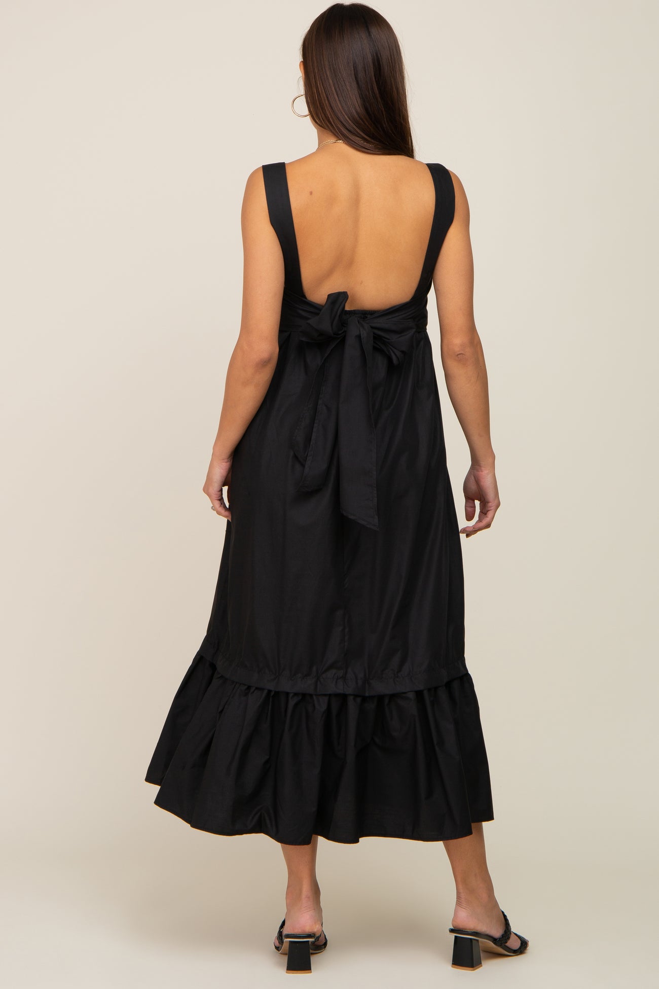 Back view of a woman wearing a black sleeveless midi dress with a bow tie back and ruffled hem.