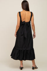 Back view of a woman wearing a black sleeveless midi dress with a bow tie back and ruffled hem.