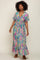 Woman with curly hair wearing a colorful floral print maxi dress with ruffle sleeves and brown sandals