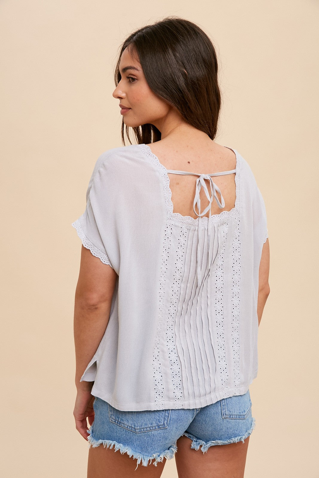 woman wearing light grey short sleeve blouse with lace trim and tie back detail paired with denim shorts
