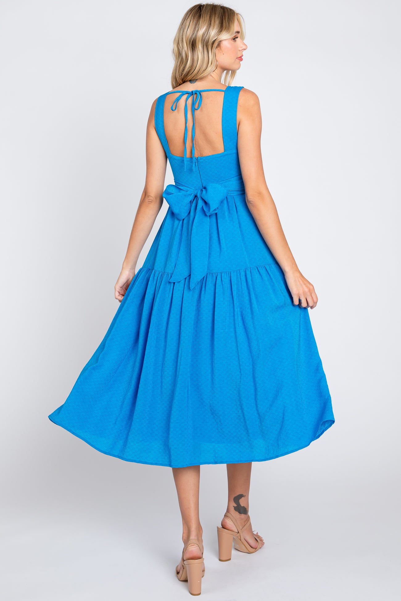 Back view of a woman wearing a bright blue midi dress with a bow tie waist and tiered skirt