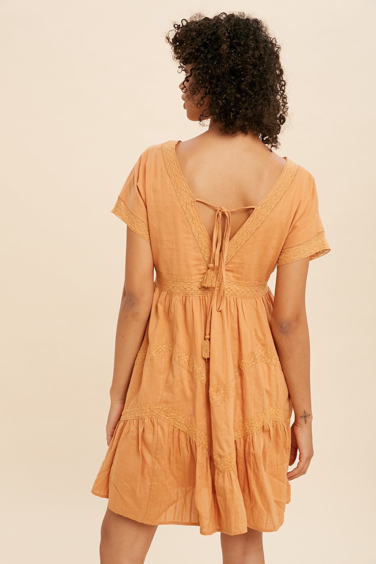 Back view of a woman with curly hair wearing an orange boho mini dress with lace trim and tassel ties