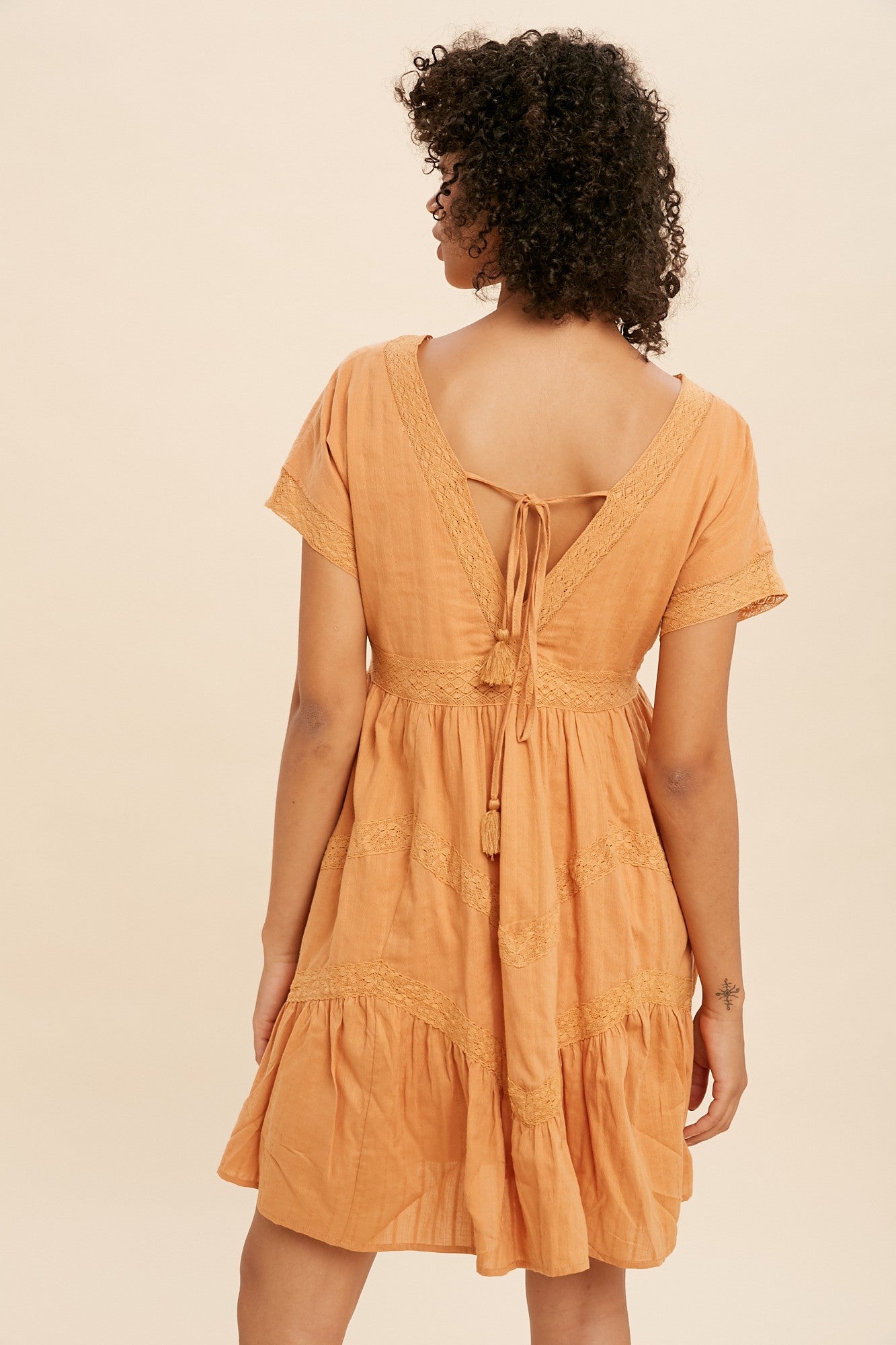 Back view of a woman with curly hair wearing an orange boho mini dress with lace trim and tassel ties