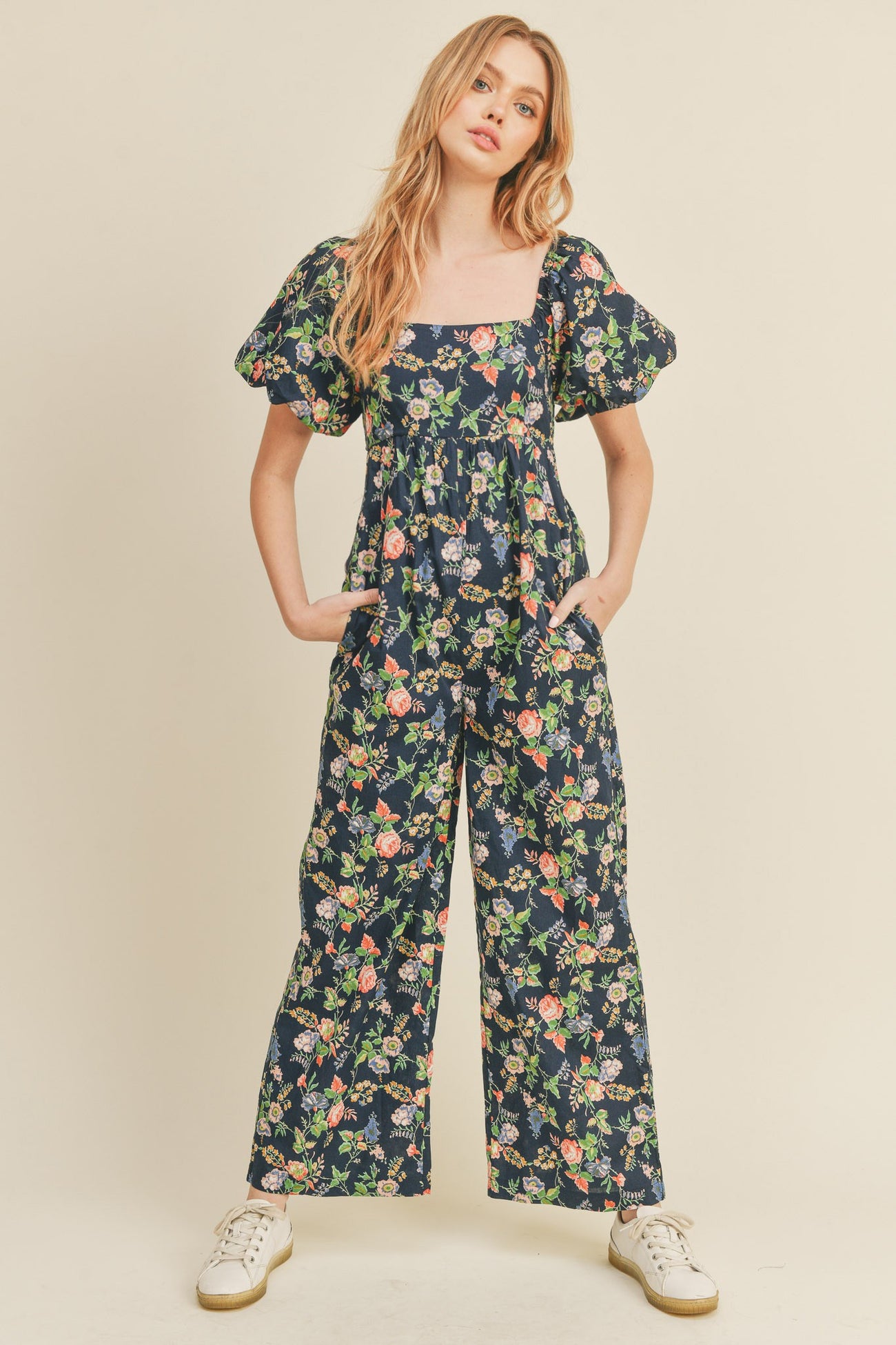 woman wearing a navy floral jumpsuit with puff sleeves and side pockets, styled with white sneakers