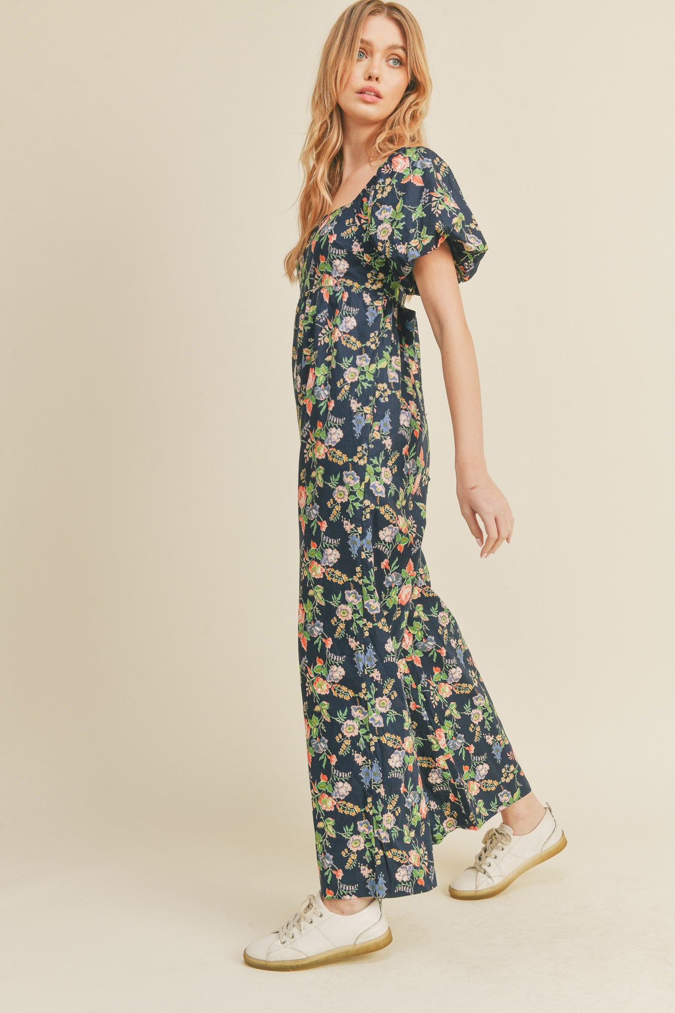 woman wearing a navy blue floral print wide leg jumpsuit and white sneakers against a neutral background