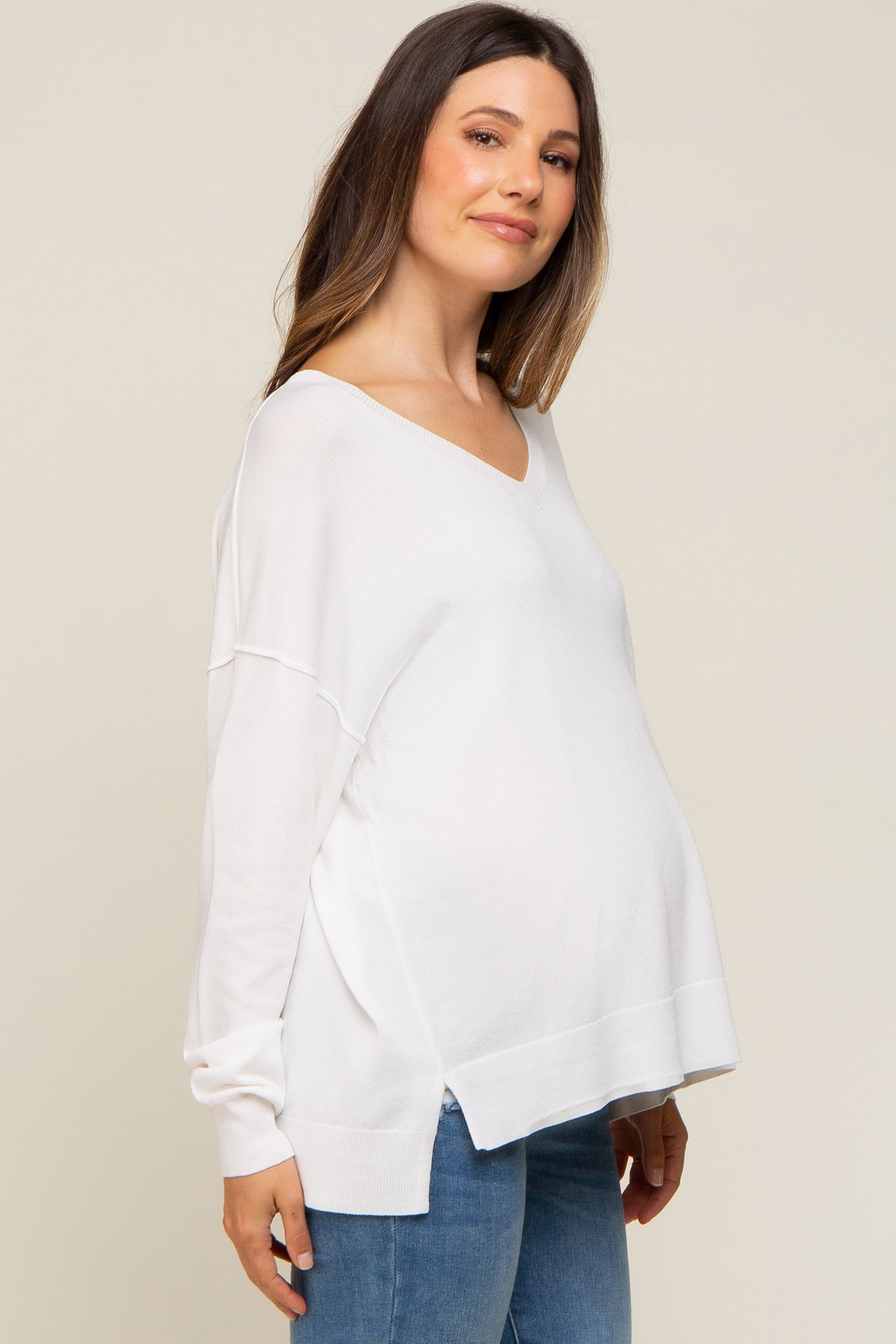 pregnant woman wearing a white v-neck maternity sweater and blue jeans against a neutral background