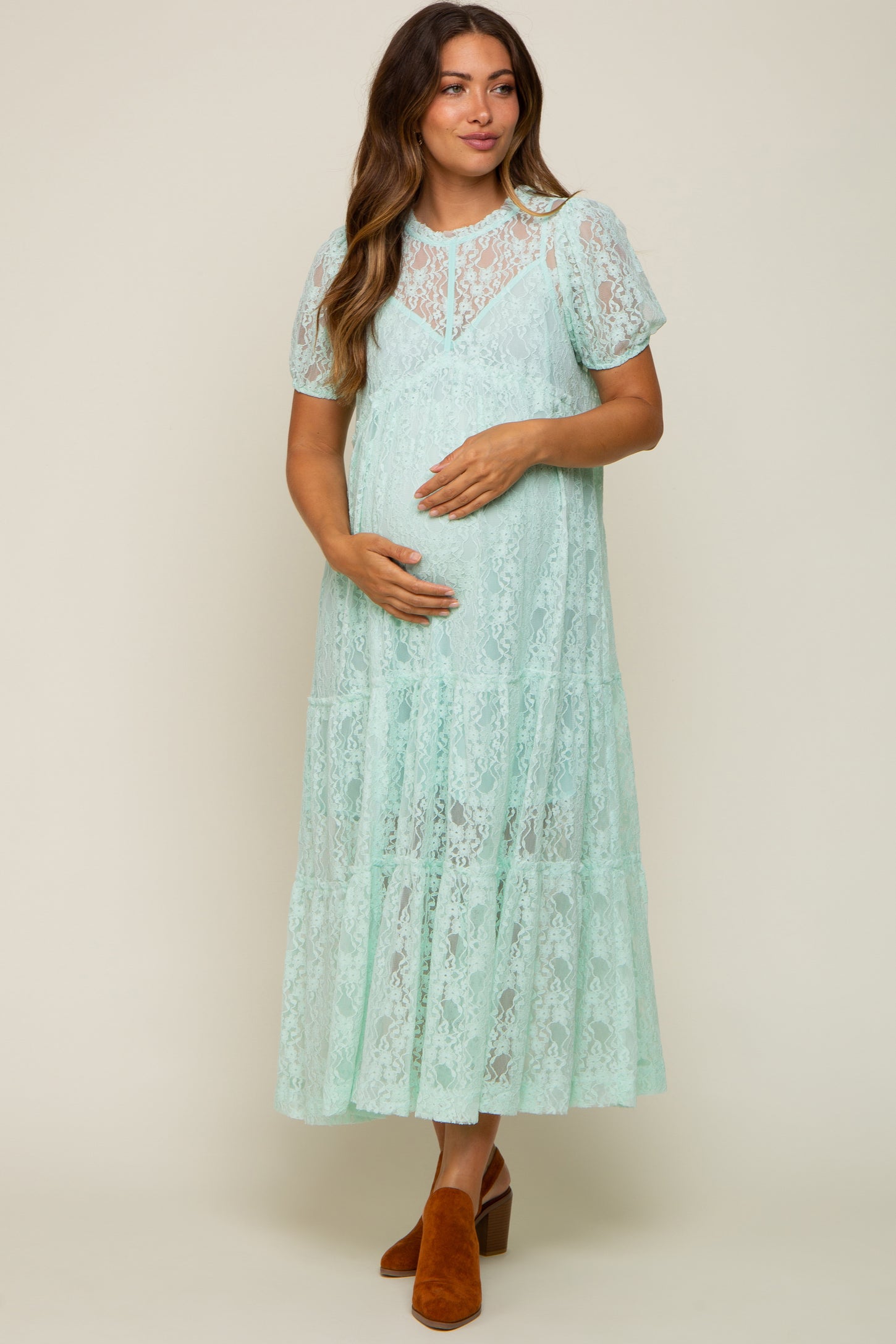 woman wearing a mint green lace maternity dress with short sleeves and a tiered skirt, cradling her baby bump