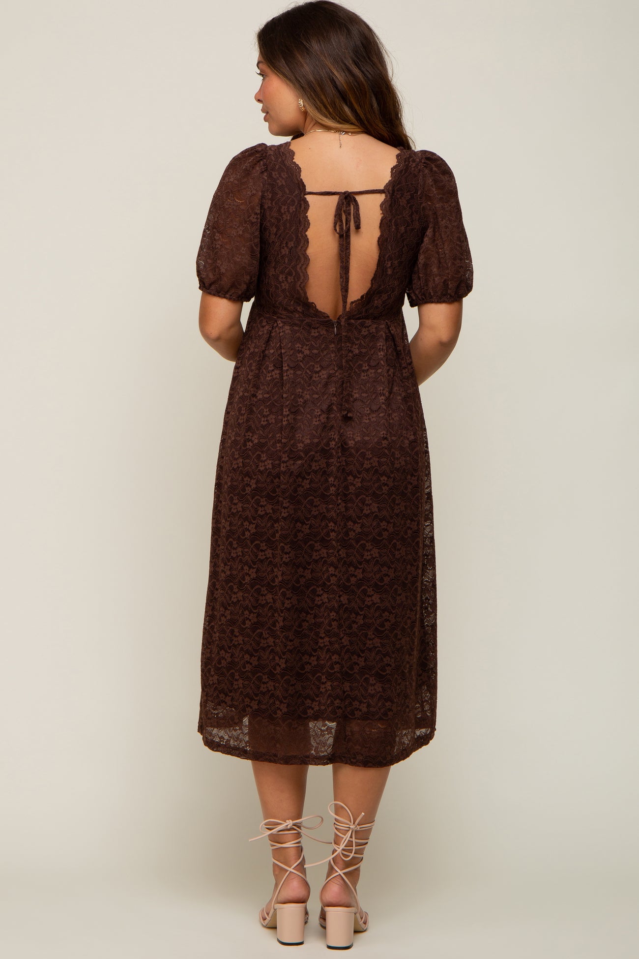 Woman wearing a brown lace midi dress with an open back and tie detail, paired with lace-up block heels.