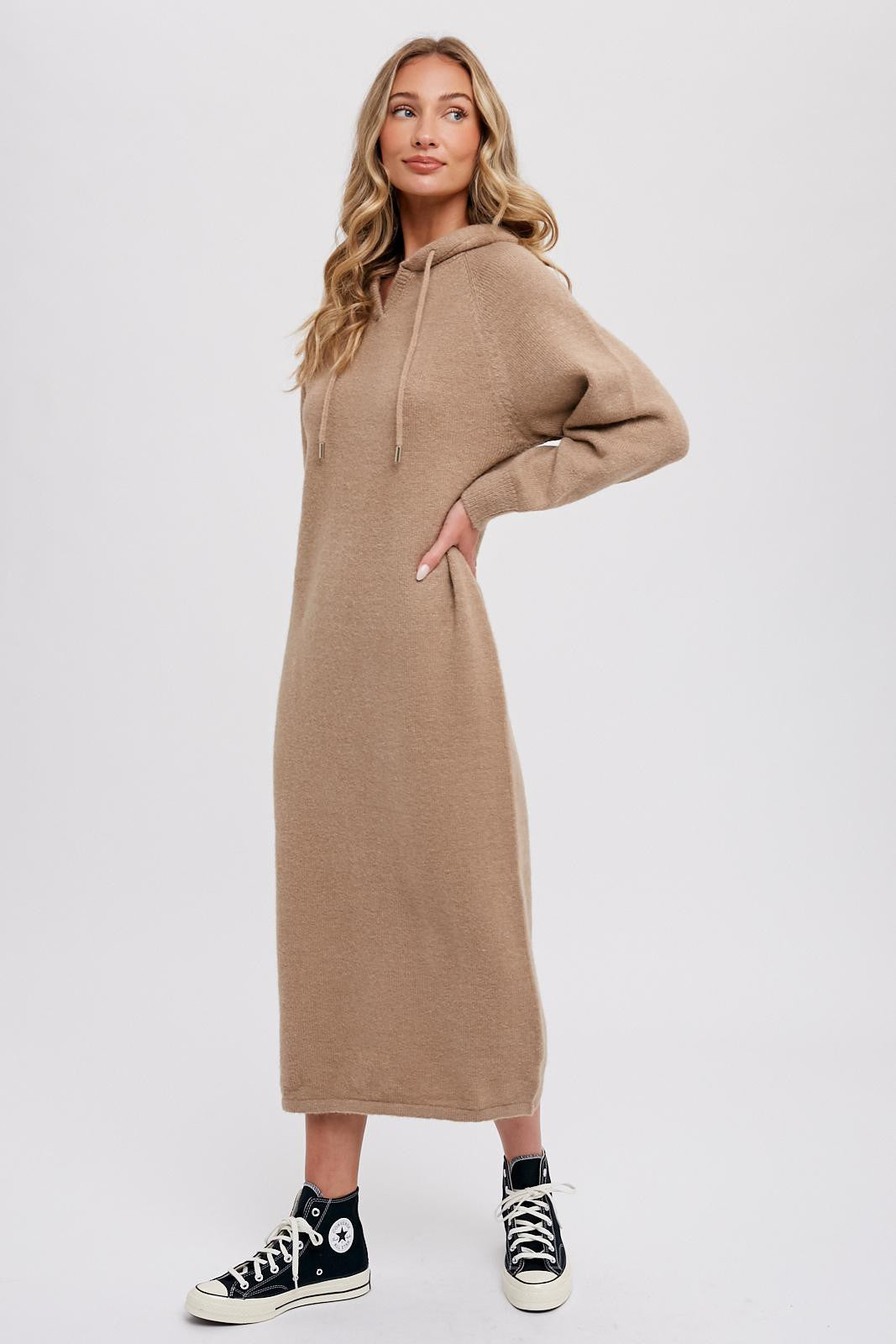 Woman wearing a tan midi-length hooded sweater dress and black high-top sneakers against a white background
