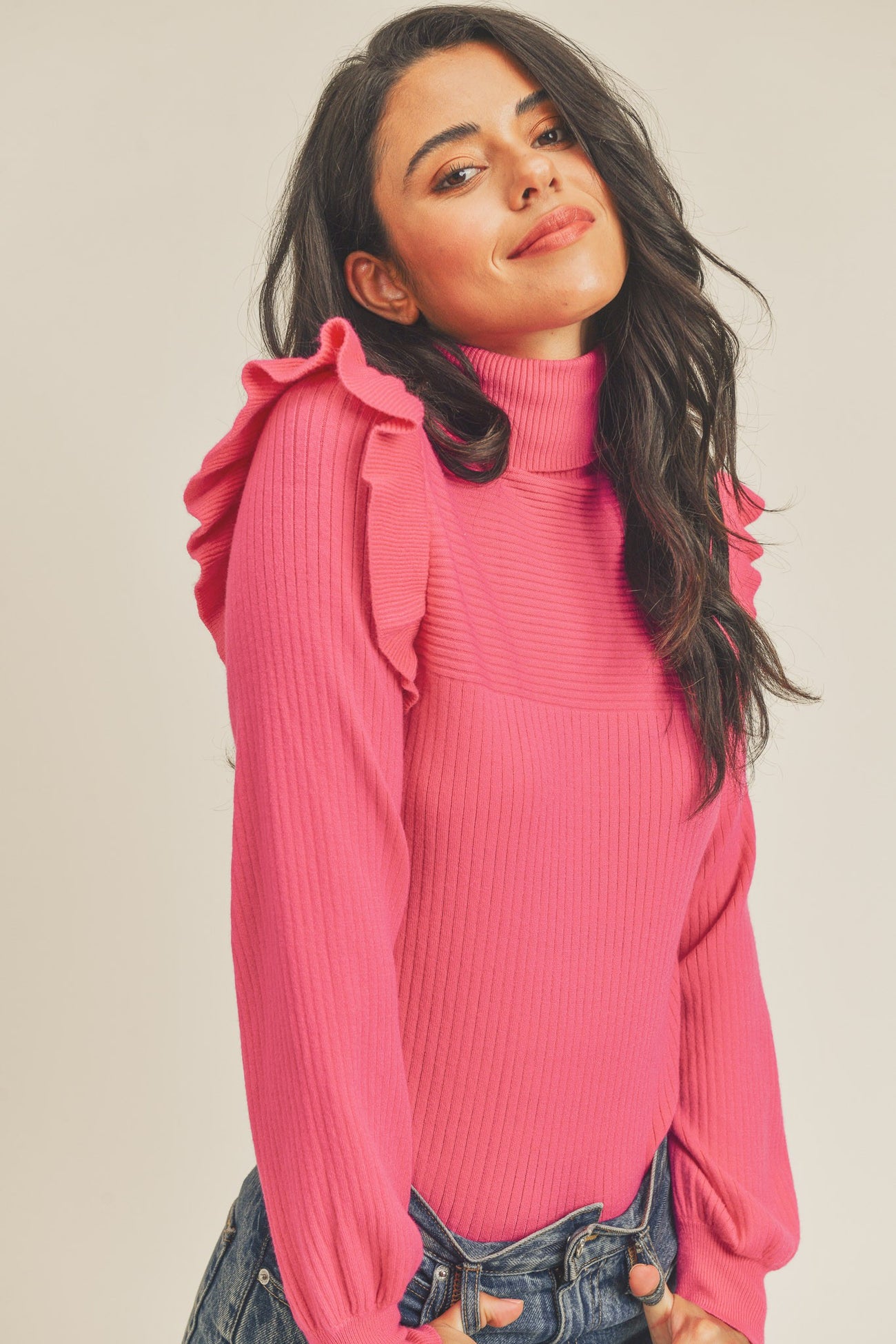 woman wearing a pink ribbed turtleneck sweater with ruffle shoulder details and blue jeans