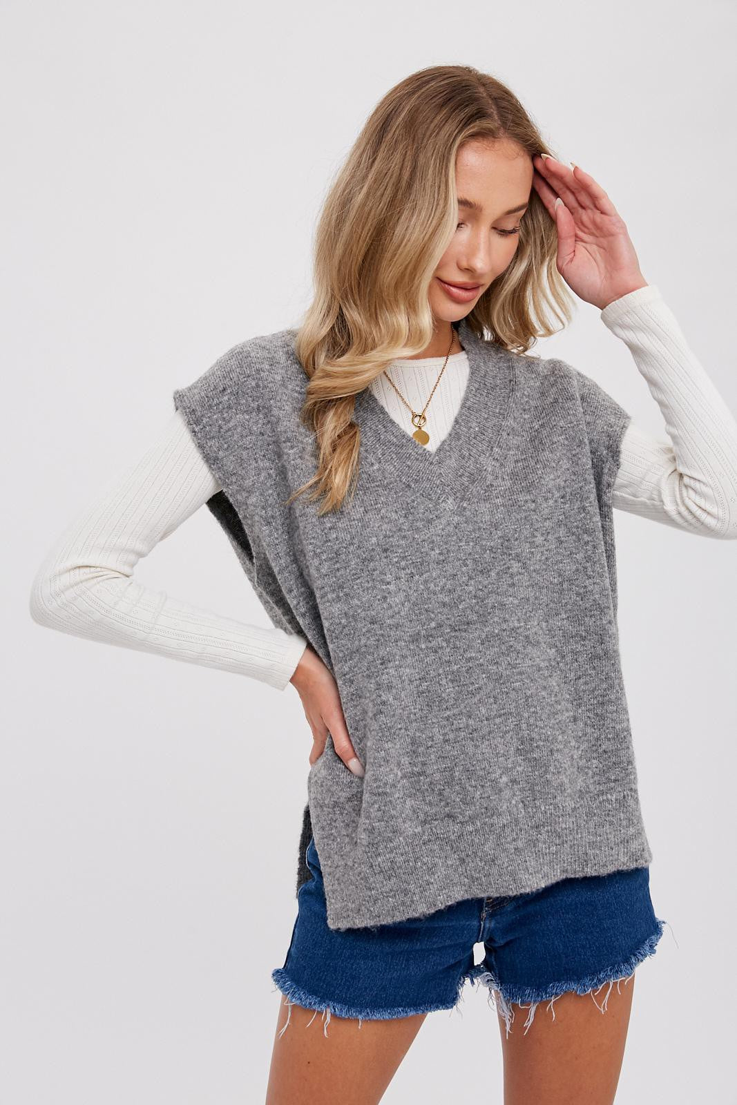 Woman wearing a grey oversized v-neck sweater vest over a white long sleeve top and denim shorts