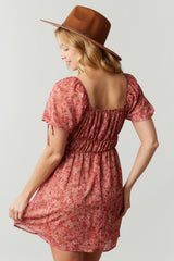 woman wearing a pink floral short dress with puff sleeves and a cinched waist, holding the skirt, and wearing a brown hat