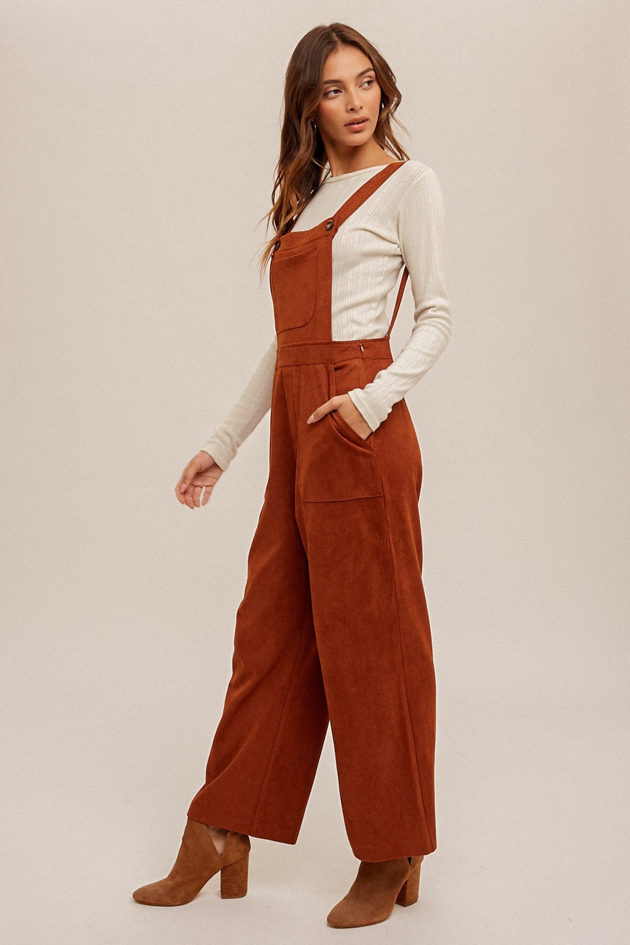 woman wearing rust-colored corduroy overalls with a white long sleeve shirt underneath and tan heeled ankle boots