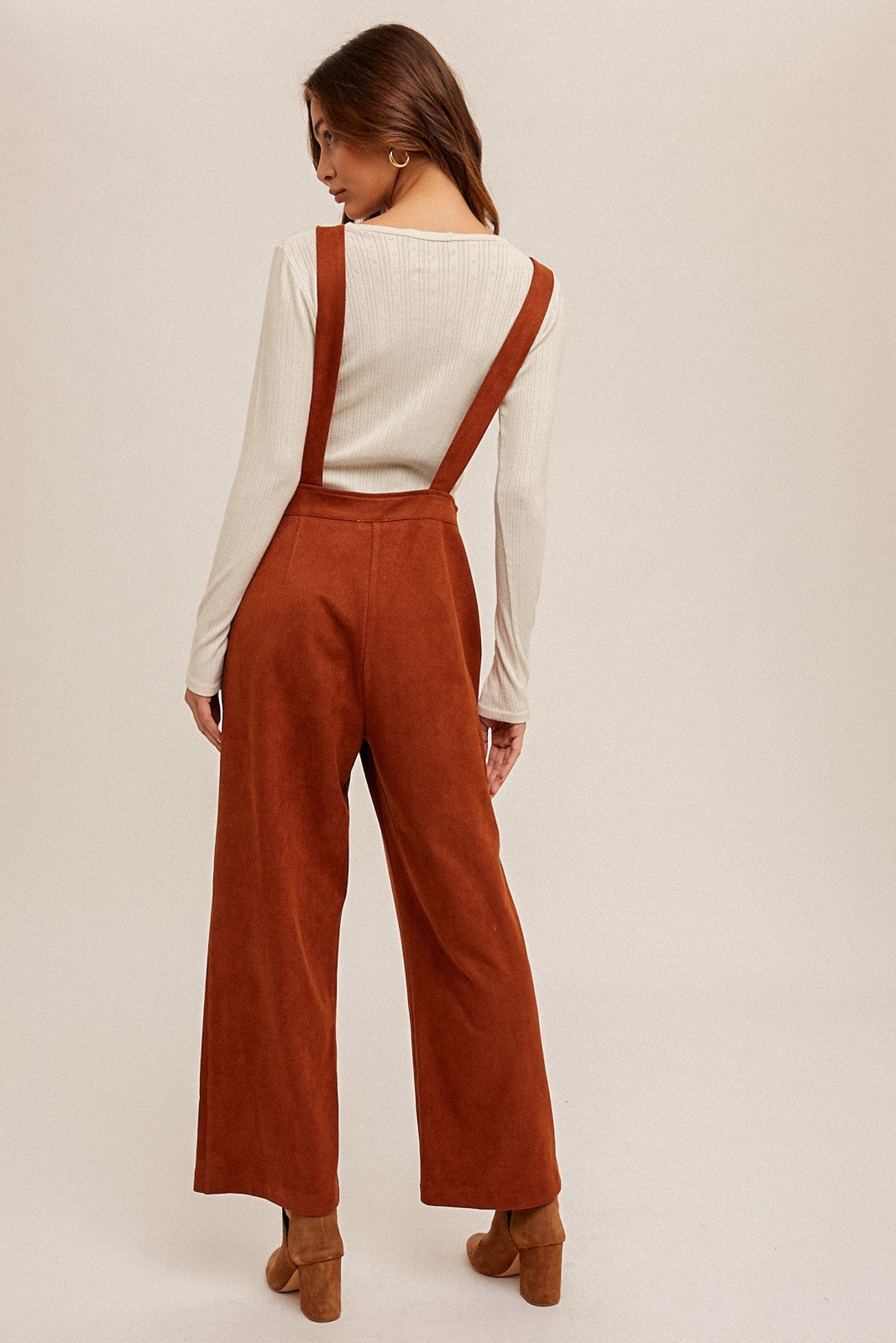 woman wearing a cream long sleeve top and rust wide leg overalls, standing with her back to the camera