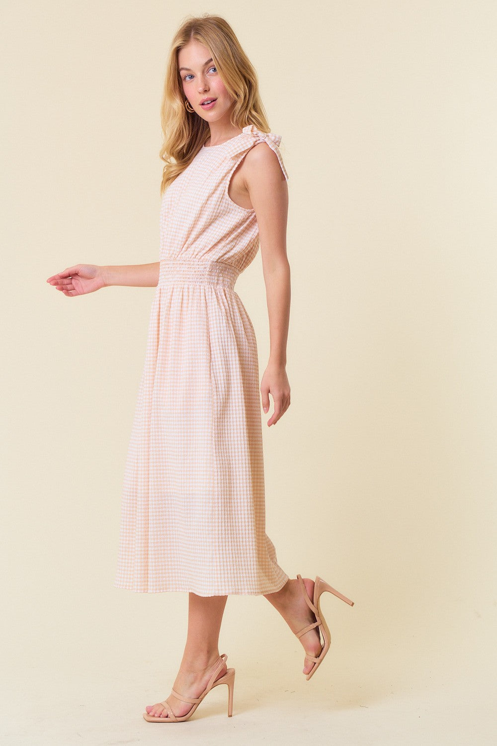 woman wearing a blush pink gingham midi dress with tie straps and an elastic waist, standing in a side pose