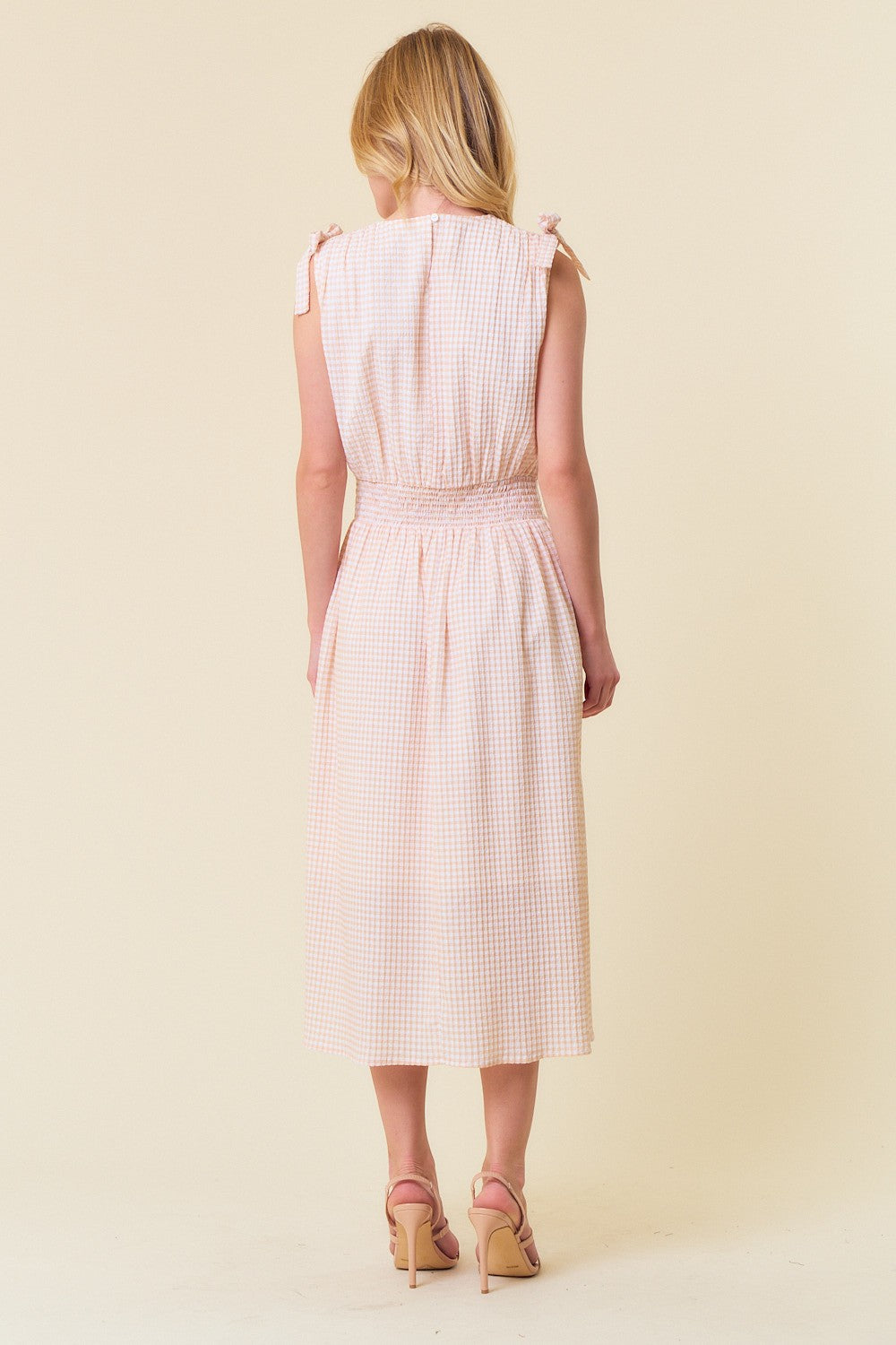 woman wearing a light pink gingham midi dress with smocked waist and tie-shoulder details, shown from the back